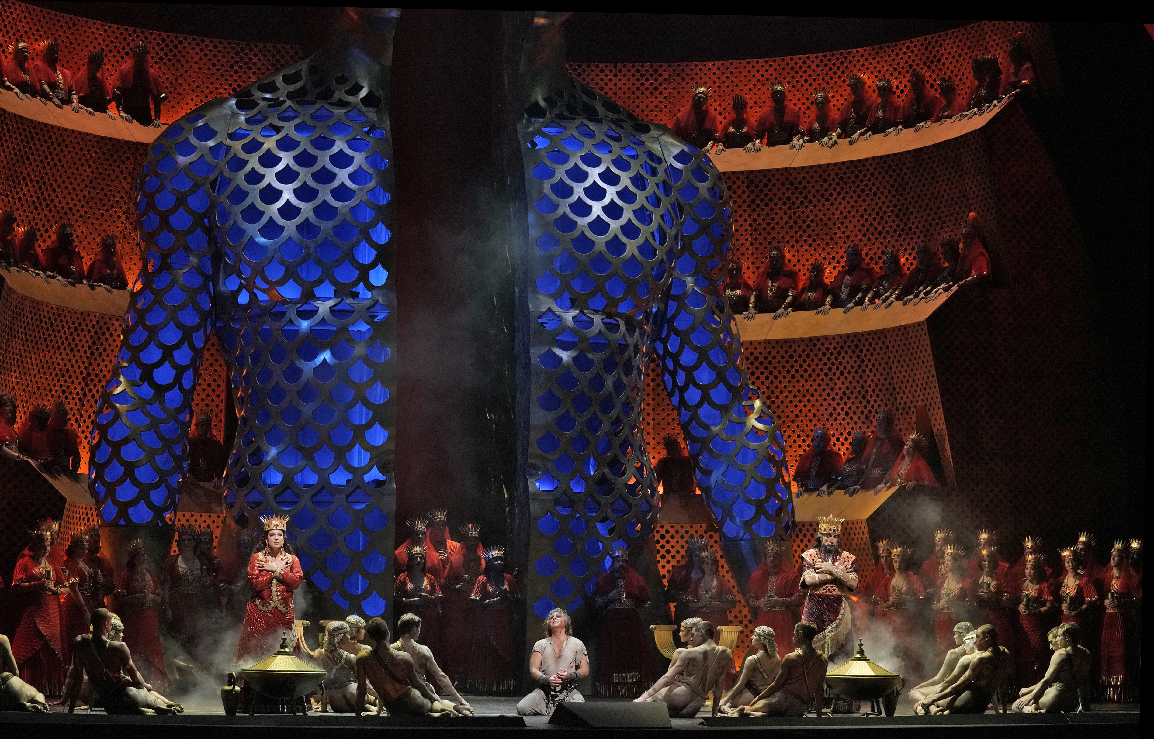 Stage scenery with a giant blue figure opening behind people sitting and praying on the floor in a circle.