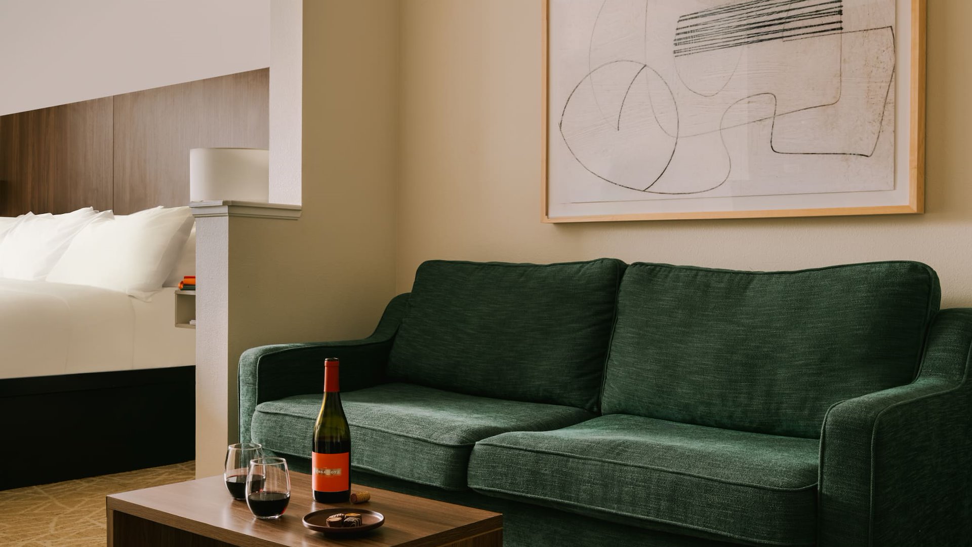 A hotel room living area with a green sofa, coffee table, and a bed in the background.