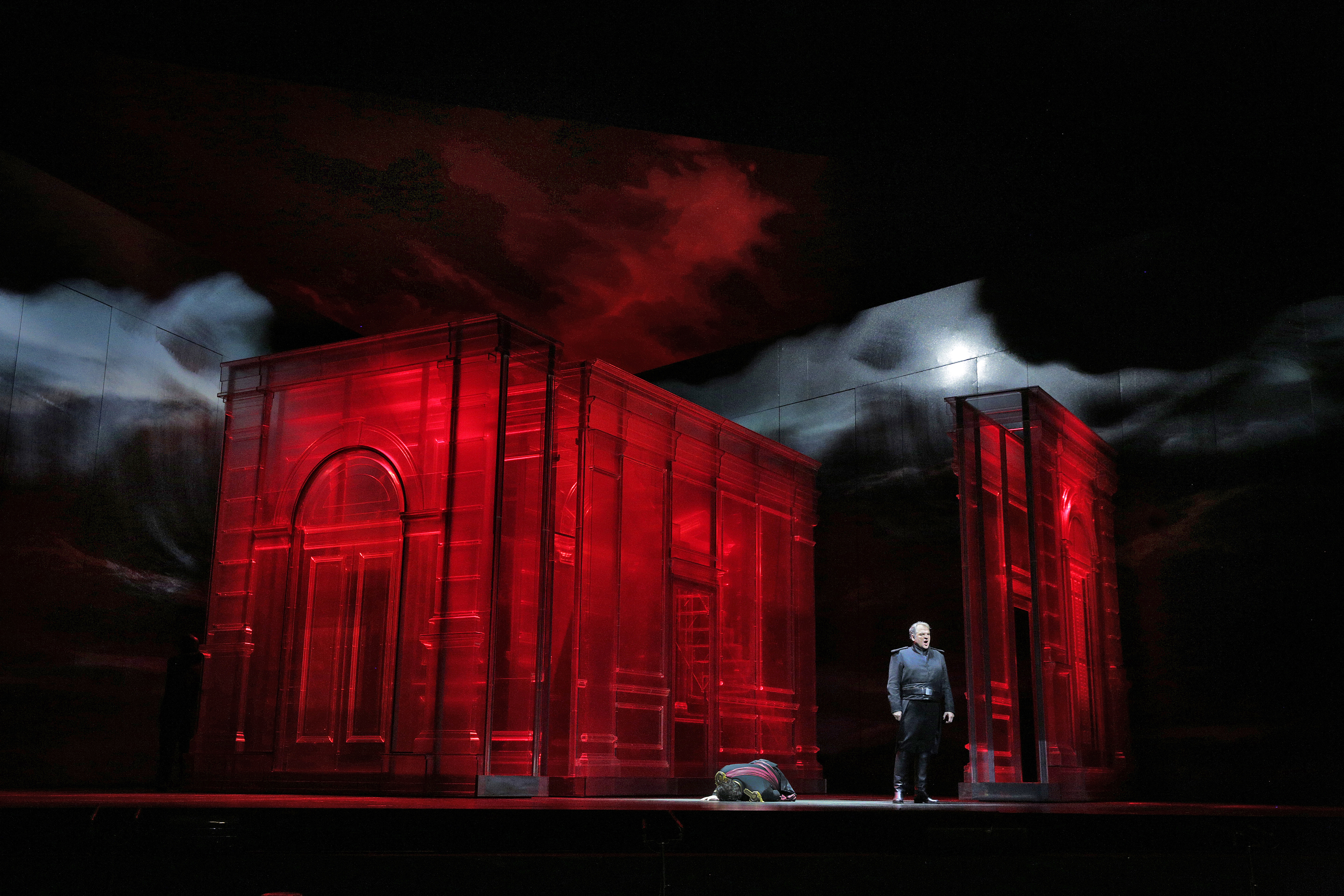 Man in a uniform singing in front of a stage scene that has a building ensconced in red, ominous-looking lighting.