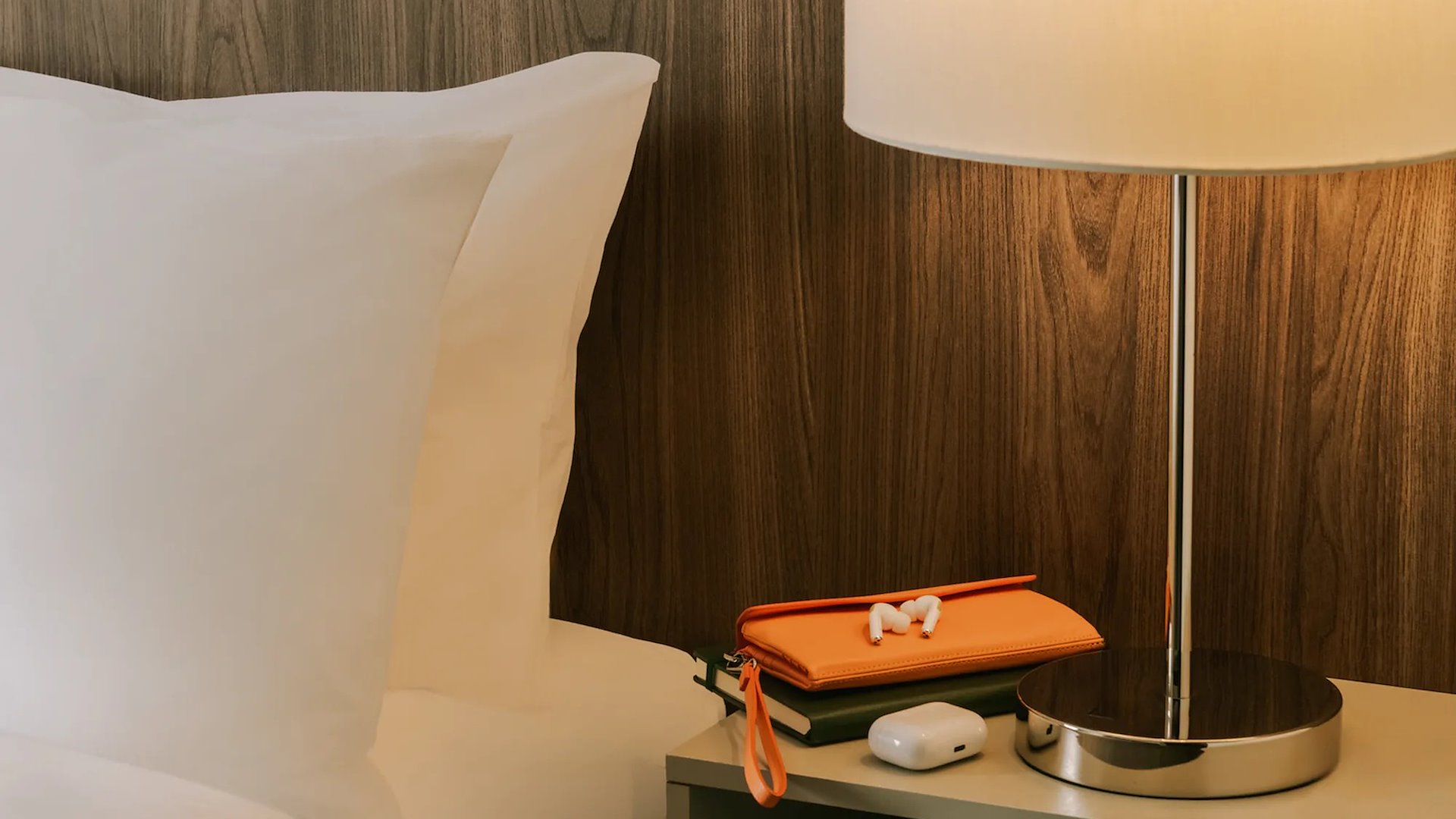 A close-up of a hotel bed with a white pillow, a lamp, and a nightstand with earbuds and a pouch.
