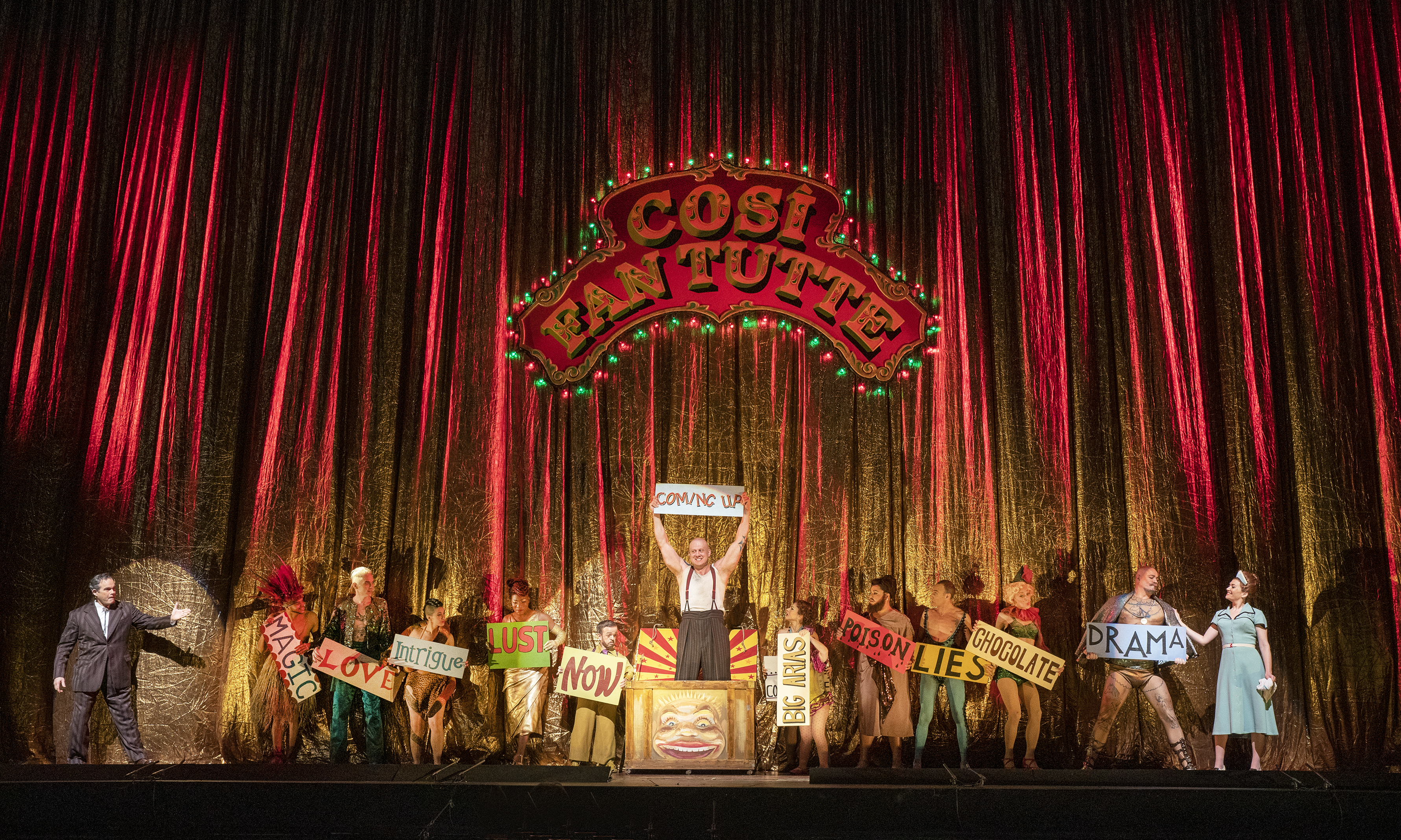 The Met Opera's cast of Manon. Man stands in a box holding a sign up while standing under sign that reads cosi fan tutte. 