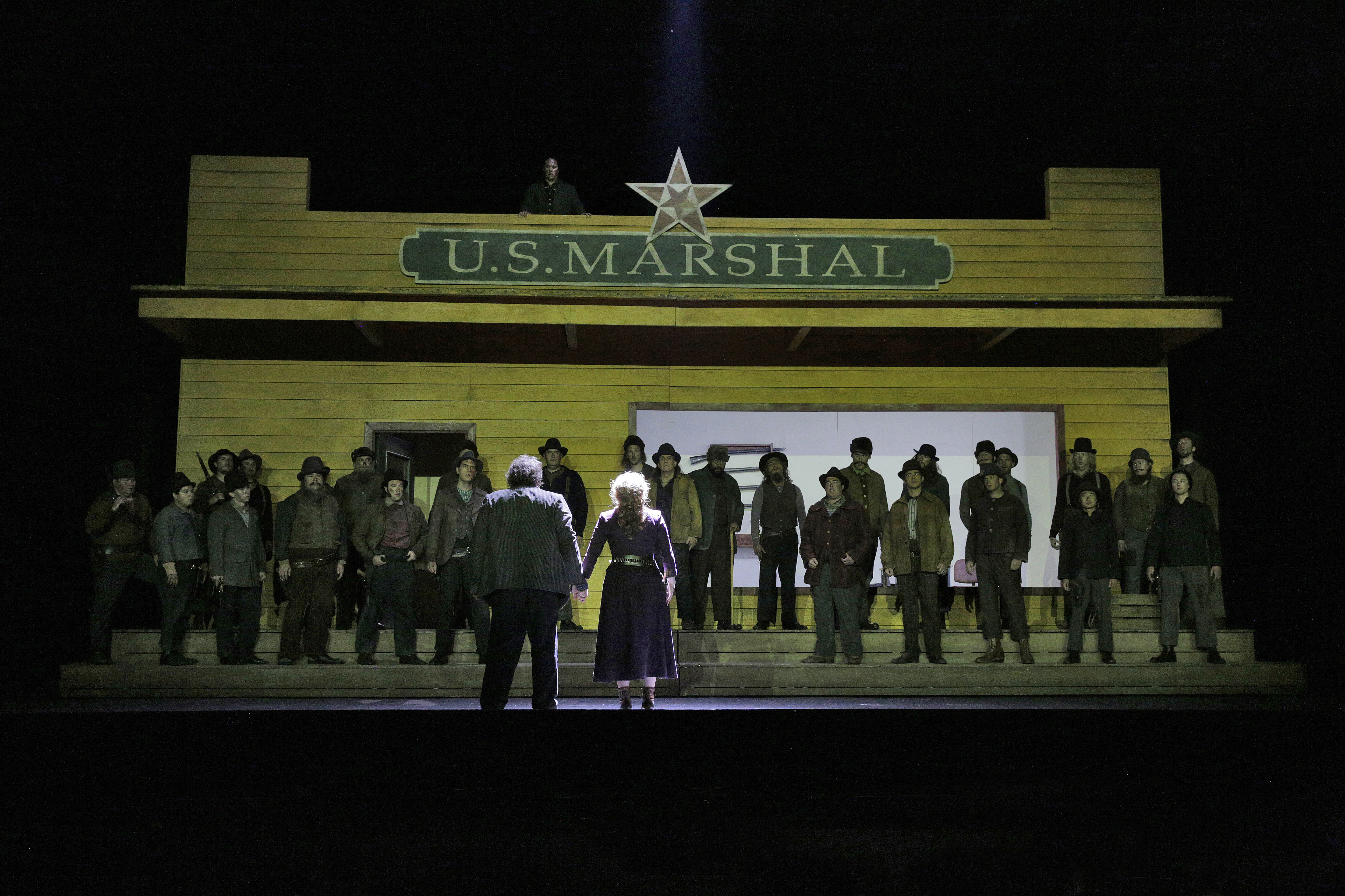 Stage scenery of a building that has "U.S. Marshal" written on it and a group of people standing and singing in front of it.