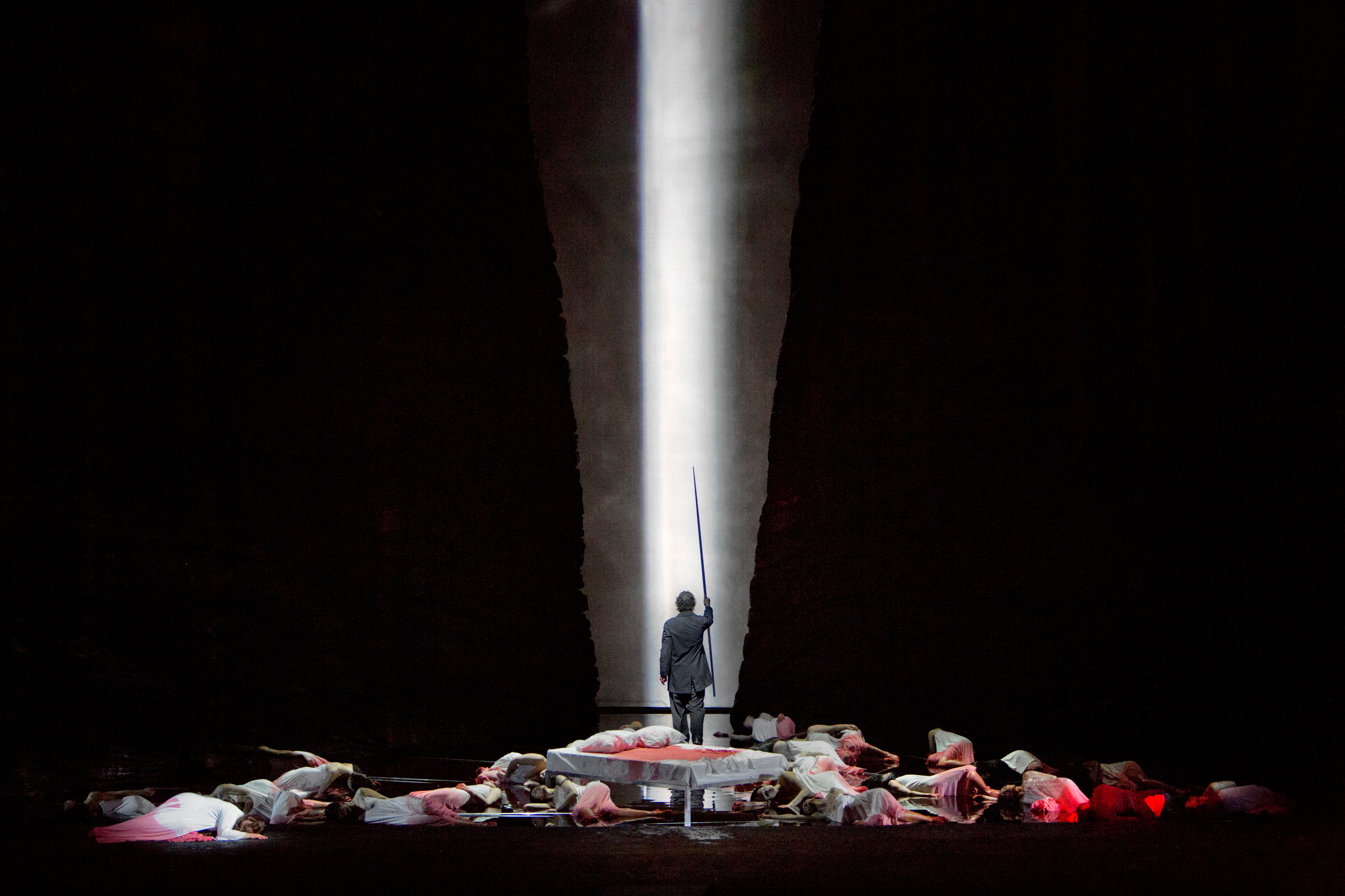 Stage depiction of multiple, fallen people looking bloodied, with a man standing behind them with his back to the audience. The man is holding a sword high in the air and is seemingly walking toward a hall or tunnel with white light coming from the other side.