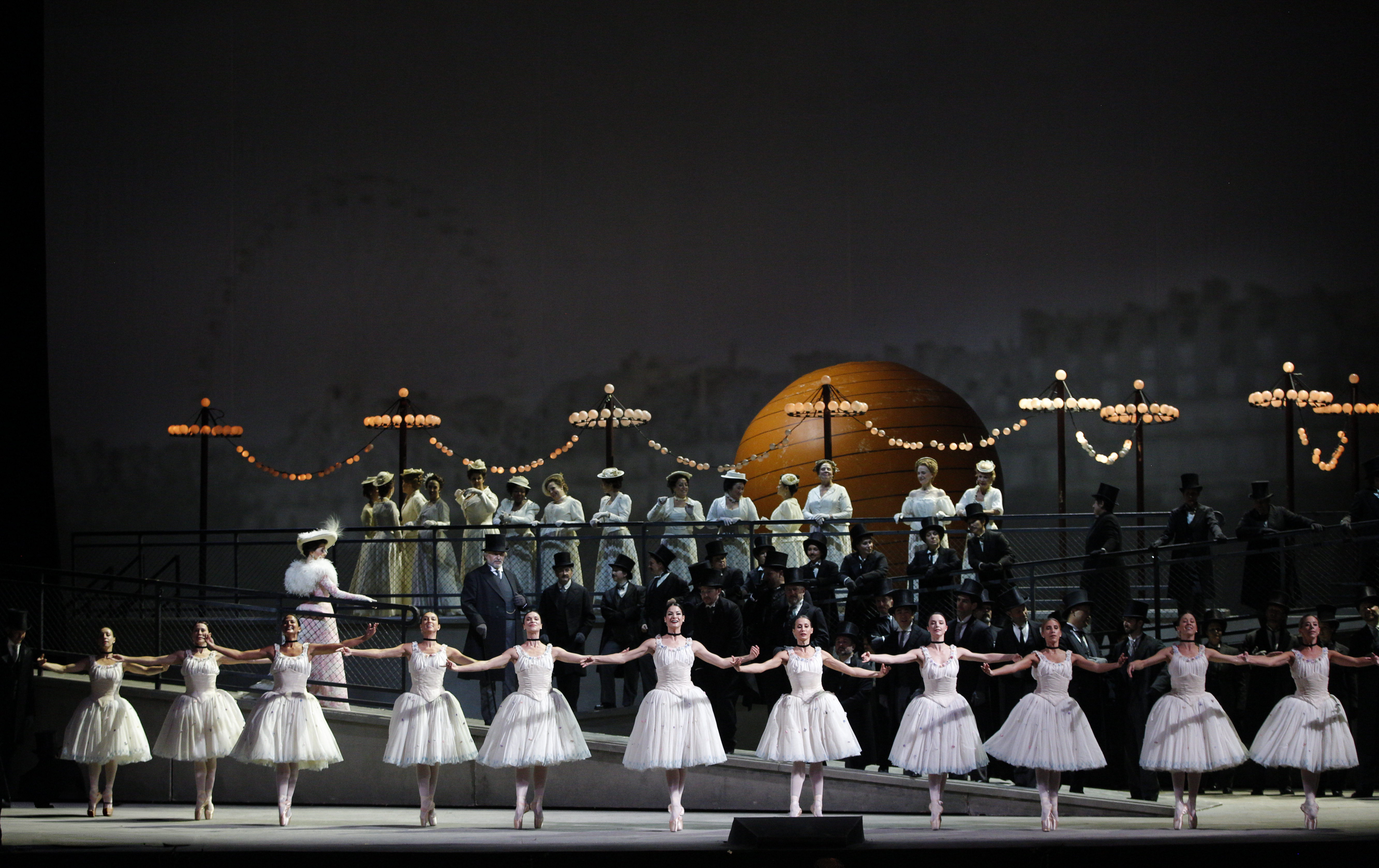 Ballet dancers on pointe in white tutus in a line, dancing in front of a stage scene of a street behind them. The street behind is filled with people in white uniforms and a woman walking by them.